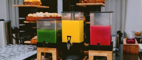 A beverage station with three dispensers, each containing a different colored drink, and a tray of pastries in the background.
