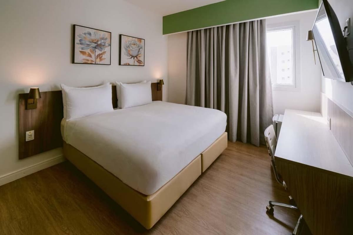 A hotel room with a large bed, a desk, a chair, two wall-mounted lamps, and a window with curtains.