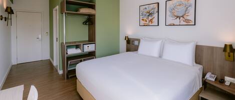 A hotel room with a large bed, a wardrobe, a desk with a chair, and two paintings on the wall.