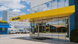 Entrada de un hotel moderno con fachada de vidrio, un toldo amarillo y un estacionamiento visible.