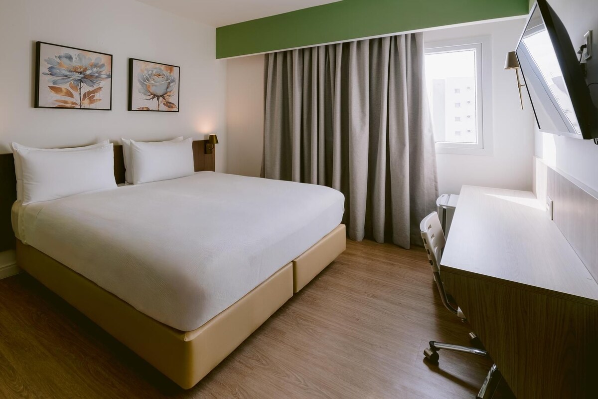 A hotel room with a large bed, a desk, a chair, a lamp, and two paintings on the wall.