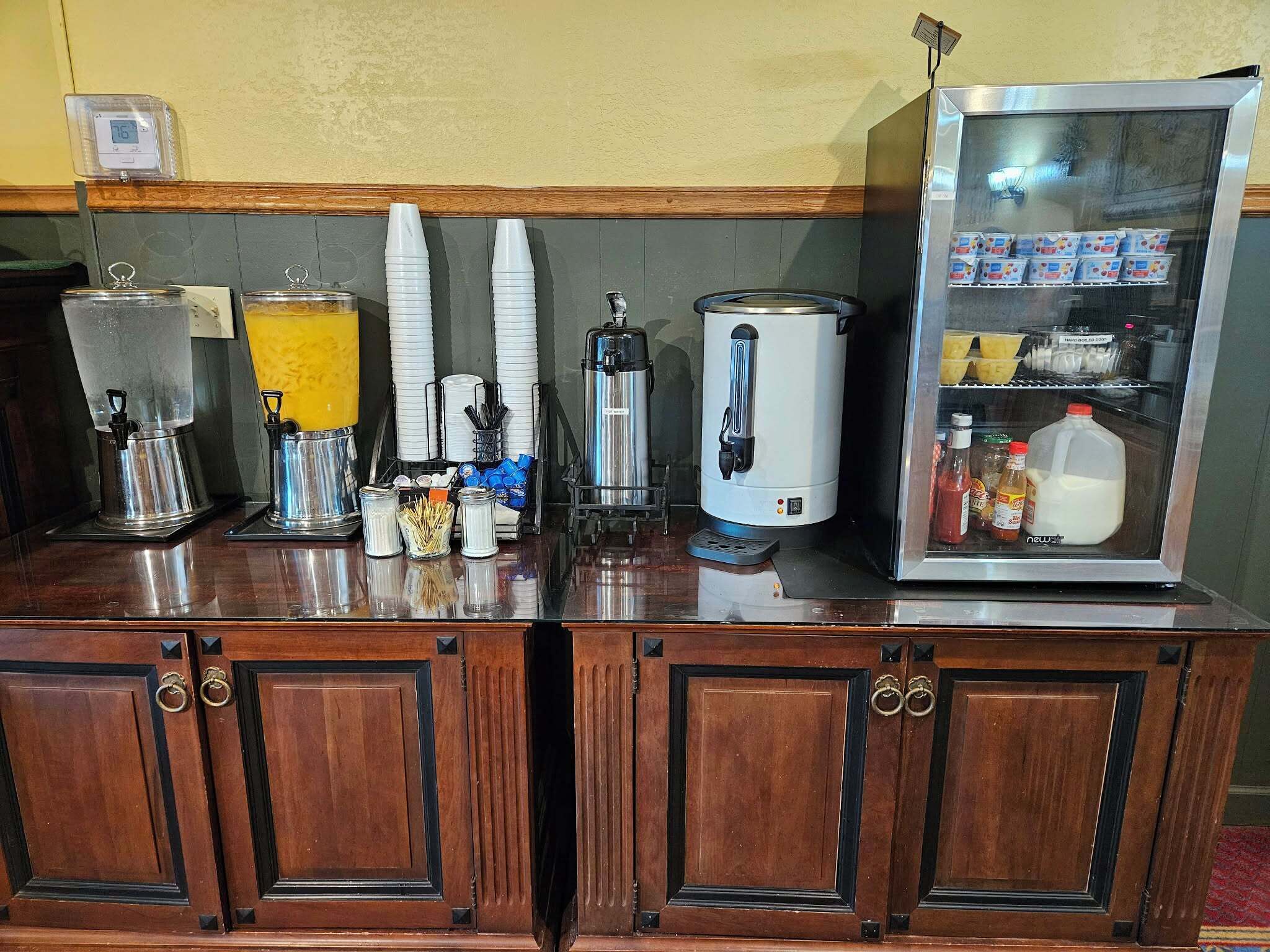 A coffee station with dispensers, cups, and a milk fridge.