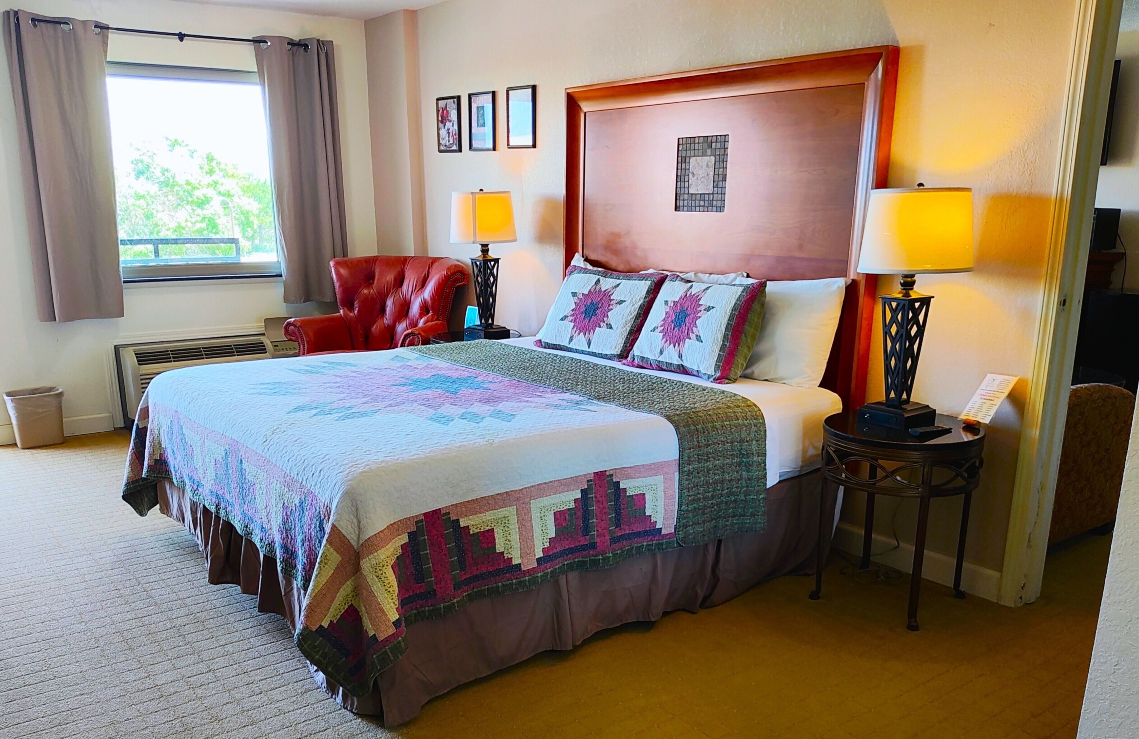 A hotel room with a large bed, two bedside lamps, a red chair, and a window with curtains.