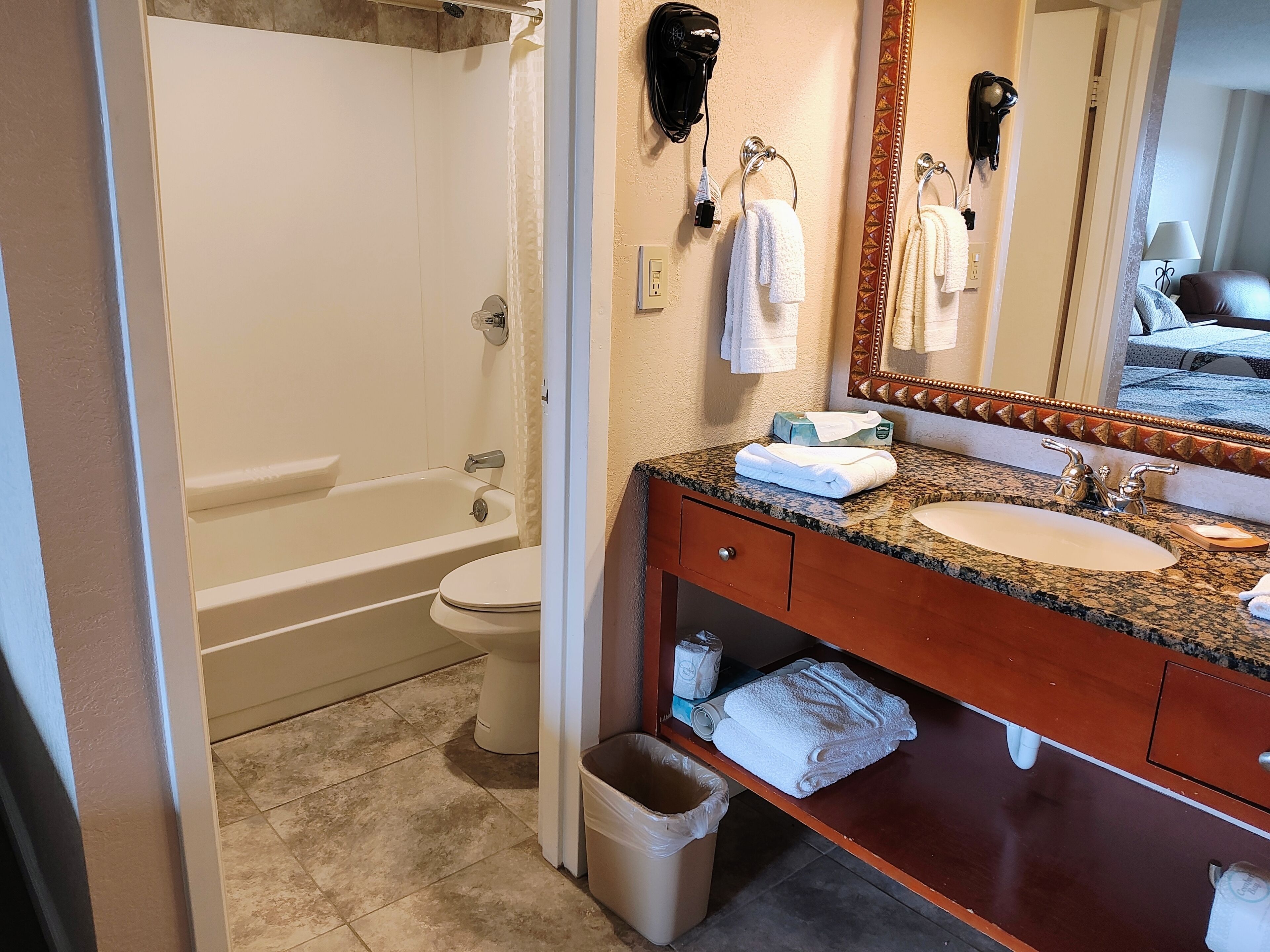 Un baño con bañera, inodoro, lavabo y tocador con espejo. Se ven toallas y un cubo de basura.