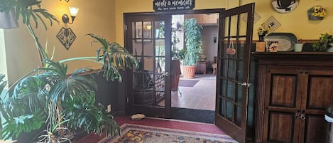 Quarto com carpete estampado, uma cadeira de madeira, um vaso de planta, uma placa na parede e uma porta que dá para o exterior.