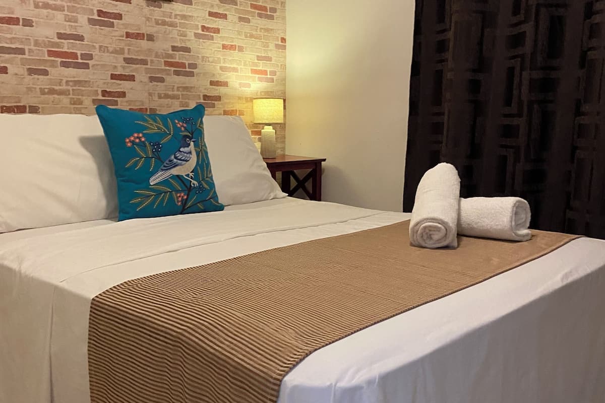 Een hotelkamer met een bed, een blauw kussen met een vogelprint, een bruine geweven plaid en een witte deken opgerold aan het voeteneinde van het bed.