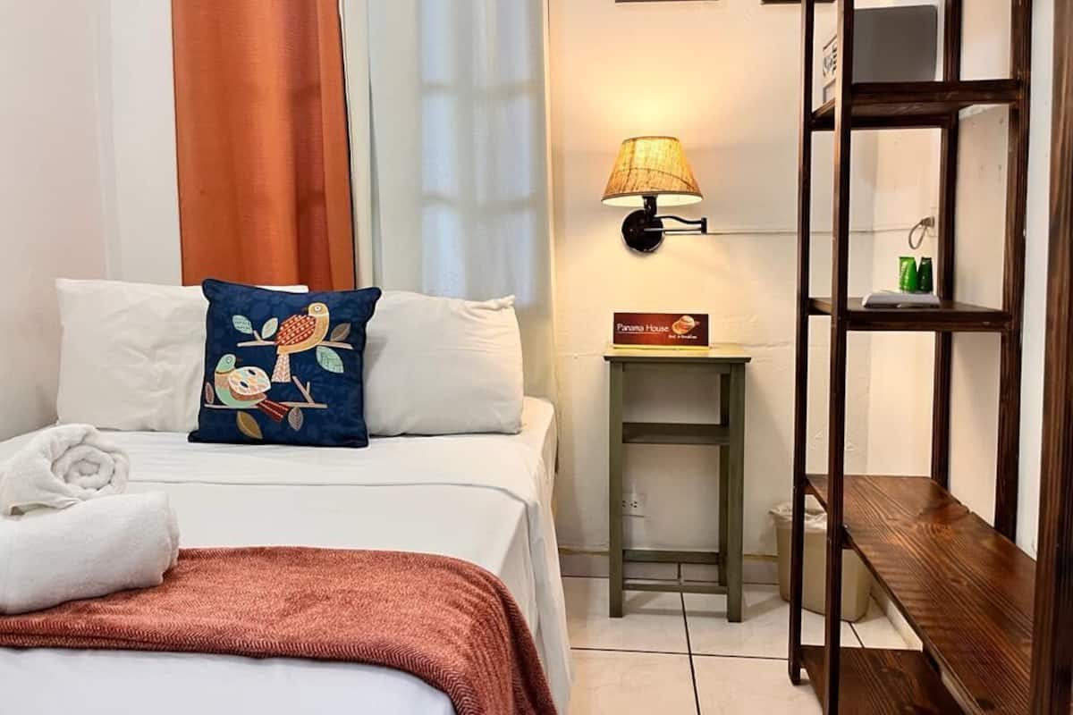 Een hotelkamer met een bed, nachtkastje, lamp, plank en een schilderij aan de muur.