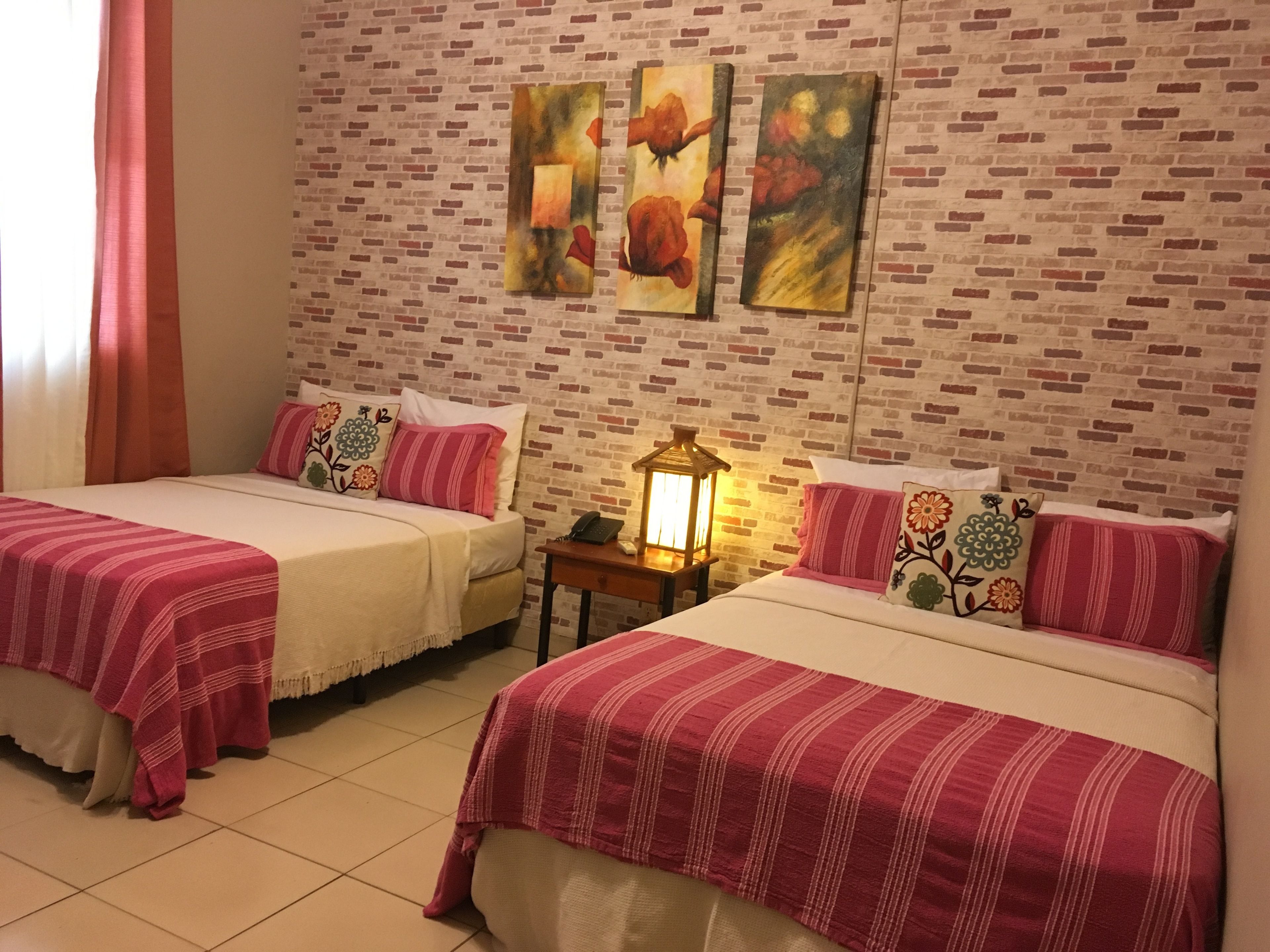 Habitación de hotel con dos camas, cada una con colcha a rayas rojas y blancas, pared de ladrillo y tres cuadros con motivos florales sobre las camas.