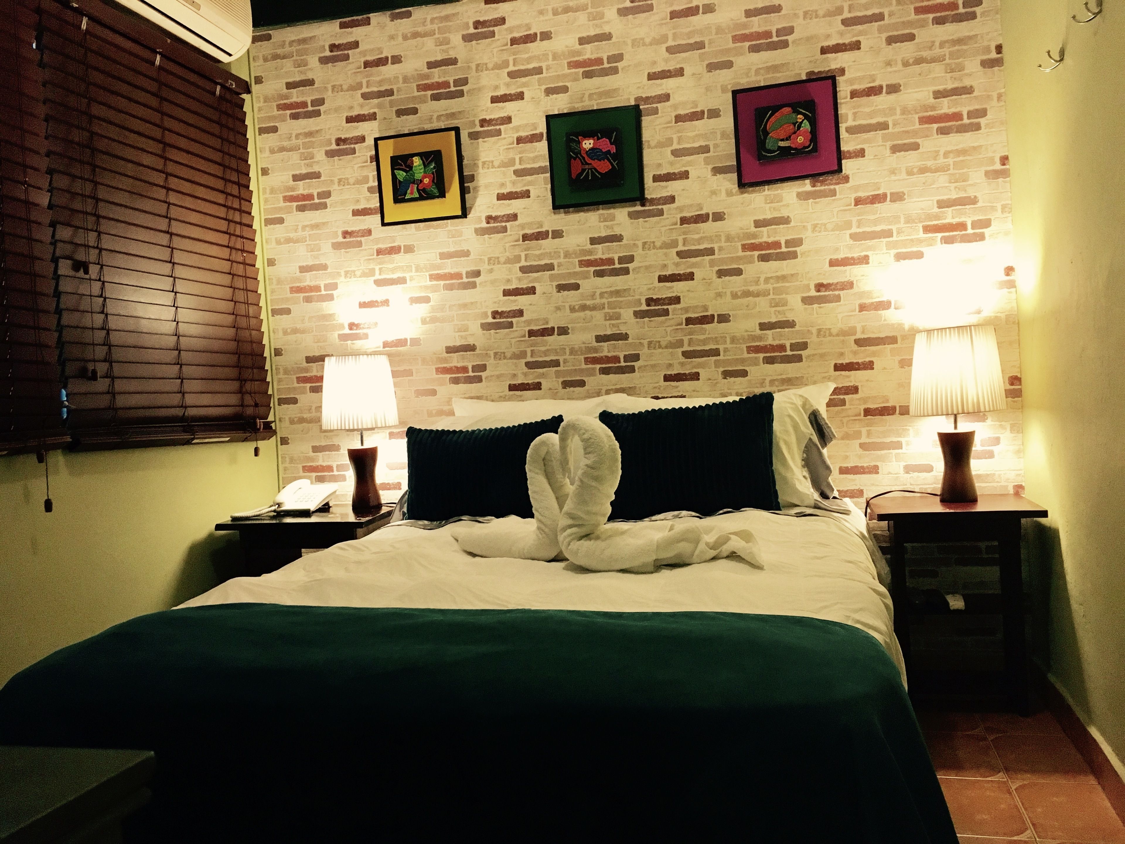 A hotel room with a brick-patterned wall, a bed with white and green bedding, two bedside tables with lamps, and a window with blinds.