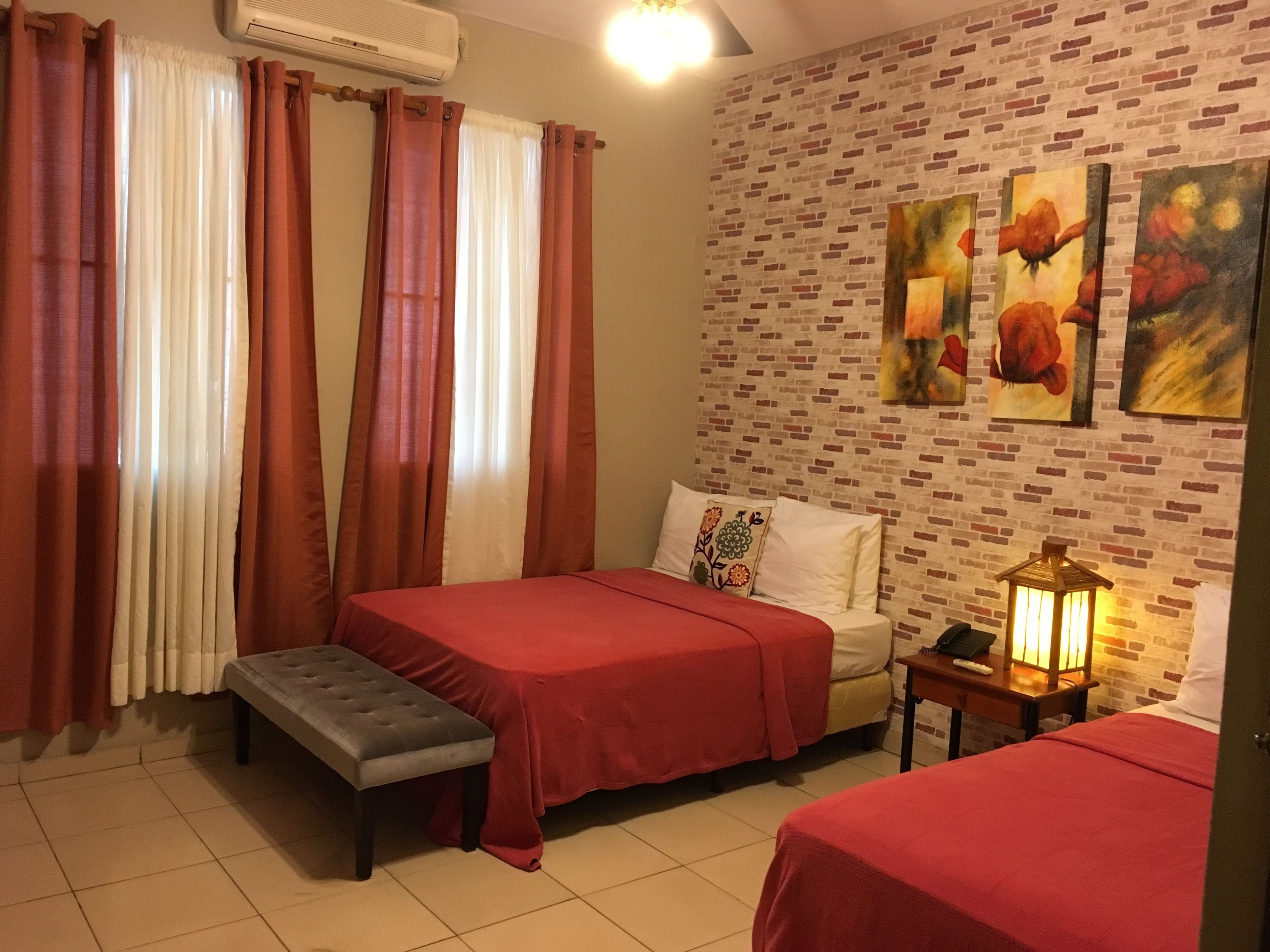 A hotel room with two beds, a bench, a bedside table with a lamp, and a brick-patterned wall with floral paintings.