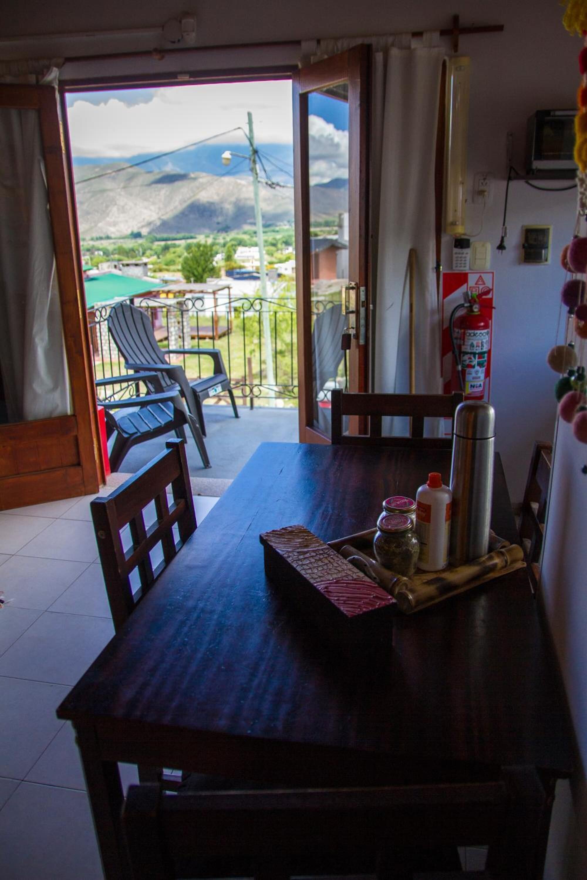 A wooden dining table with chairs, a fire extinguisher, and a view of mountains through an open door.