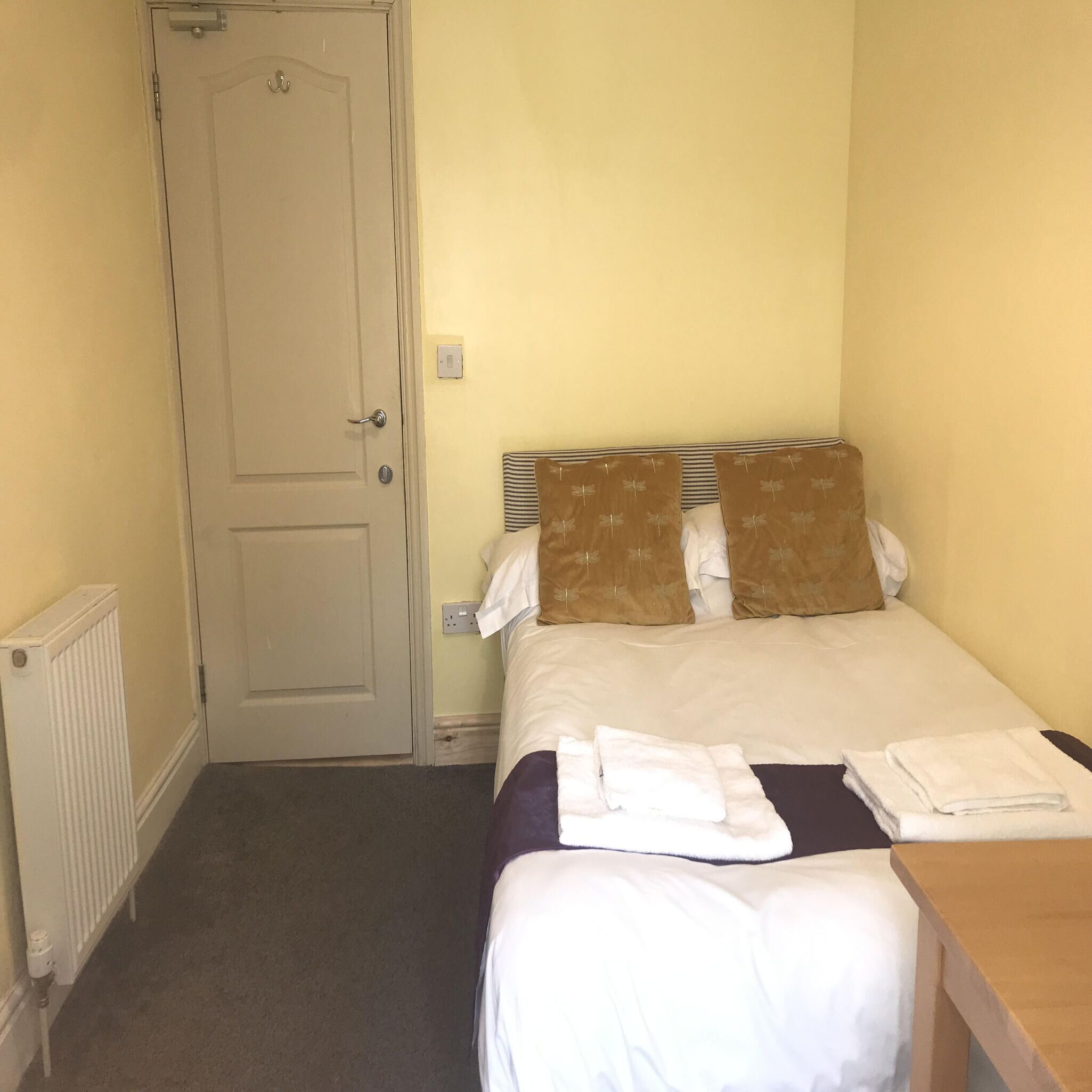 A small, single-bed room with a plain door, a radiator, and a bedside table with towels.