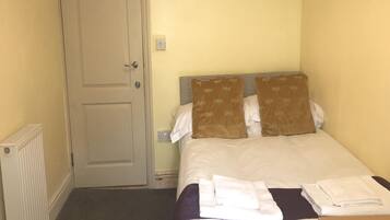 A small, single-bed room with a plain door, a radiator, and a bedside table with towels.