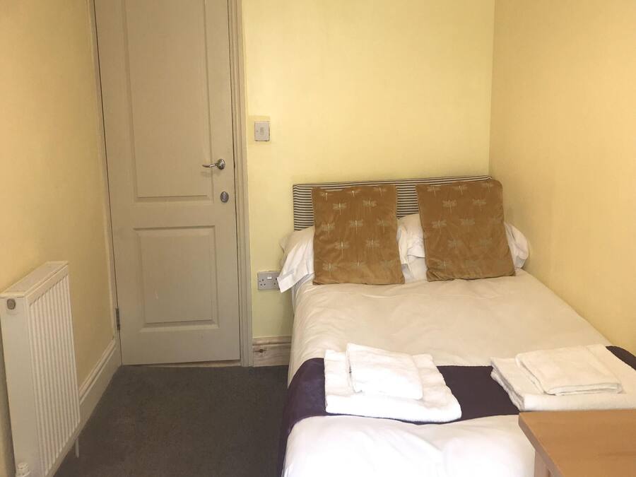 A small, single-bed room with a plain door, a radiator, and a bedside table with towels.