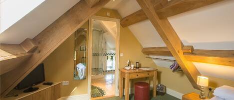 A bedroom with a sloped ceiling, wooden beams, a bed, a bedside table, a lamp, and a view into an adjacent bathroom.