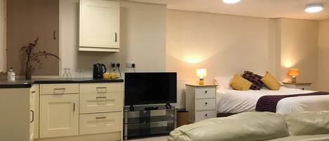 Una habitación de hotel con kitchenette, televisión, cama con almohadas y sofá.