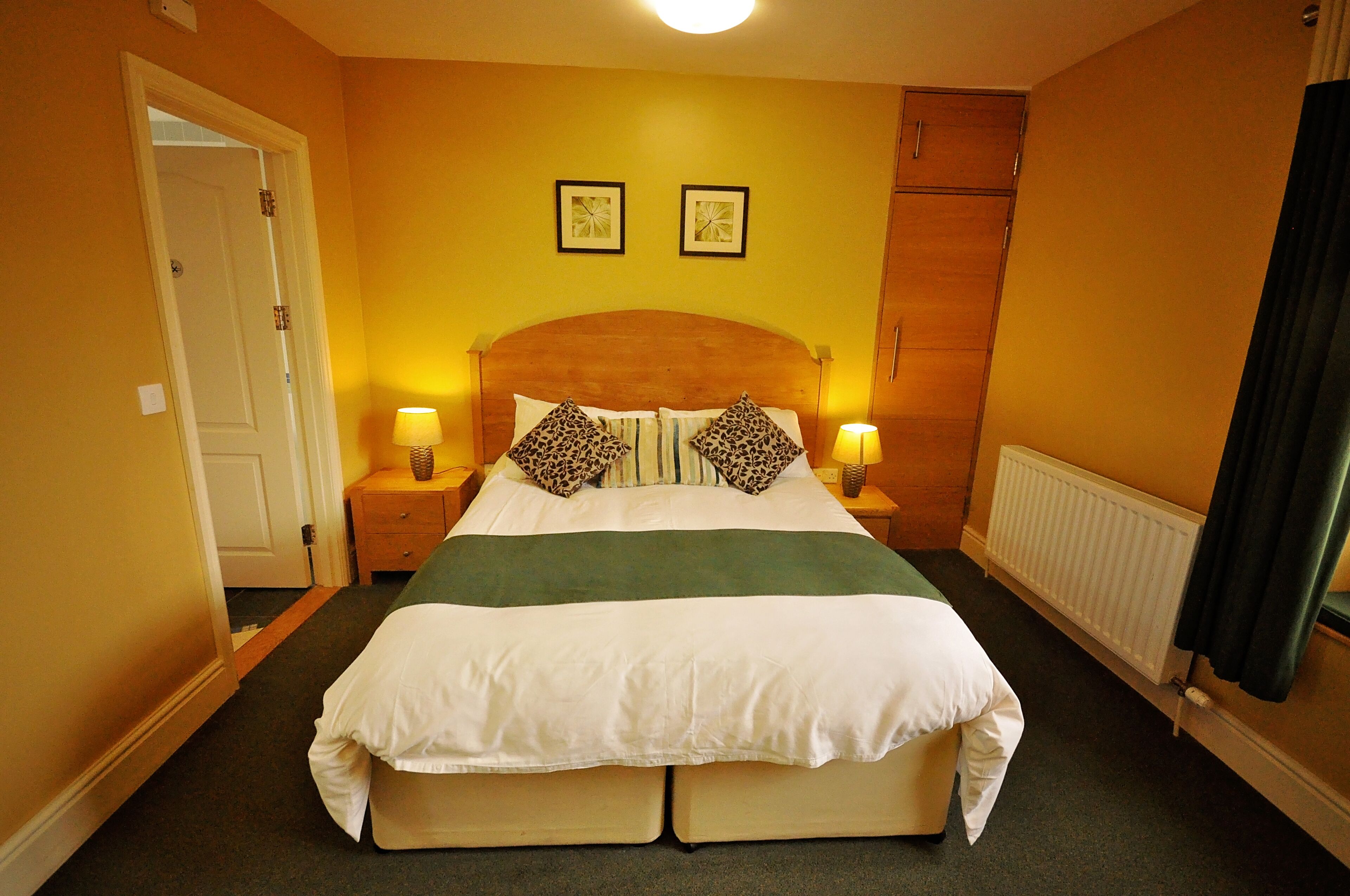 Une chambre d’hôtel avec un grand lit, deux lampes de chevet, une tête de lit en bois, un plaid vert et deux tableaux encadrés accrochés au mur.