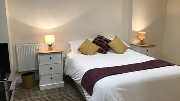 Un dormitorio ordenado con cama, dos mesitas de noche con lámparas, un radiador y un cubo de basura.