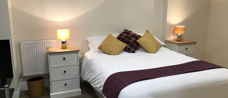 Un dormitorio ordenado con cama, dos mesitas de noche con lámparas, un radiador y un cubo de basura.