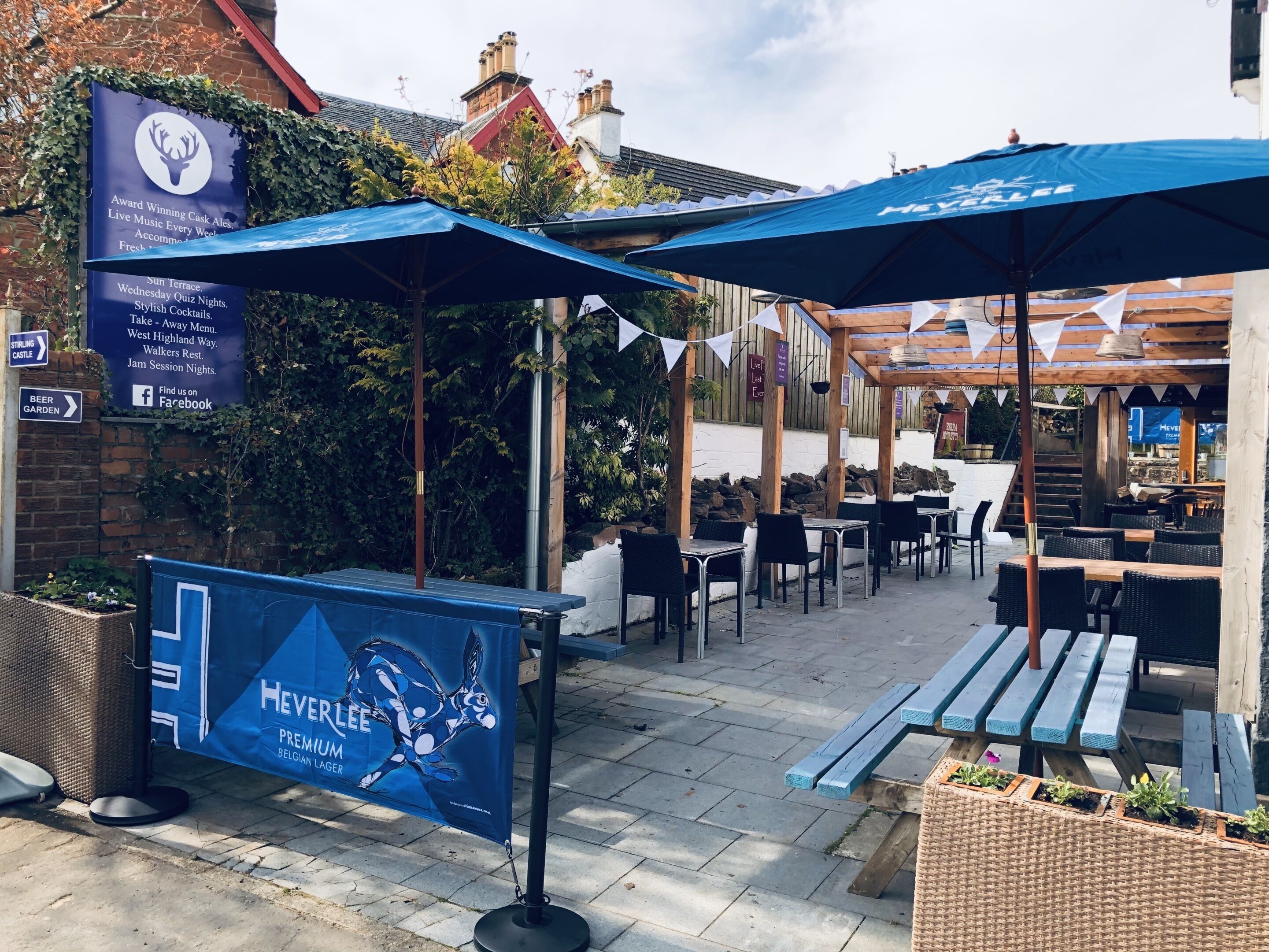 An outdoor seating area with tables and umbrellas, a banner advertising Hevelee Belgian Lager, and directional signs for nearby attractions.