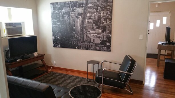 A living room with a large black and white cityscape painting, a flat-screen TV on a wooden stand, a leather sofa, a round coffee table, and a modern chair. - The Happy Hollow (Hot Springs)