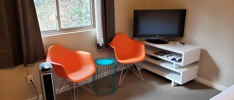 Een kamer met een televisie op een standaard, twee oranje stoelen, een kleine blauwe tafel en een witte plank met spullen.