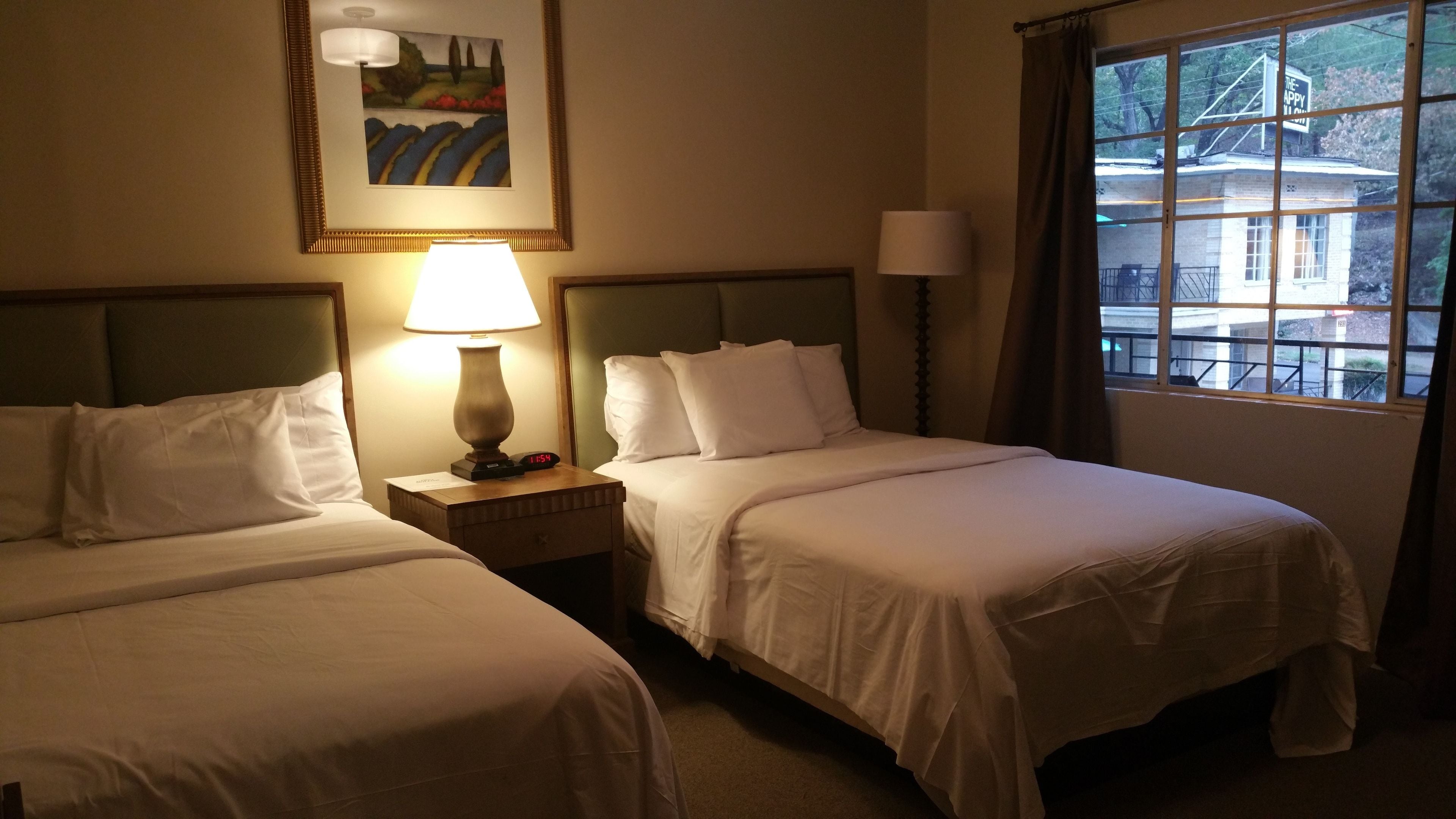 A hotel room with two single beds, a nightstand with a lamp, a painting on the wall, and a window with a view of a building.