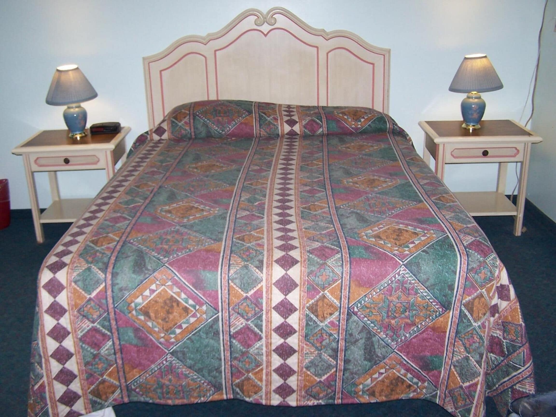 A bed with a patterned bedspread, two bedside tables with lamps, and a nightstand with a lamp.