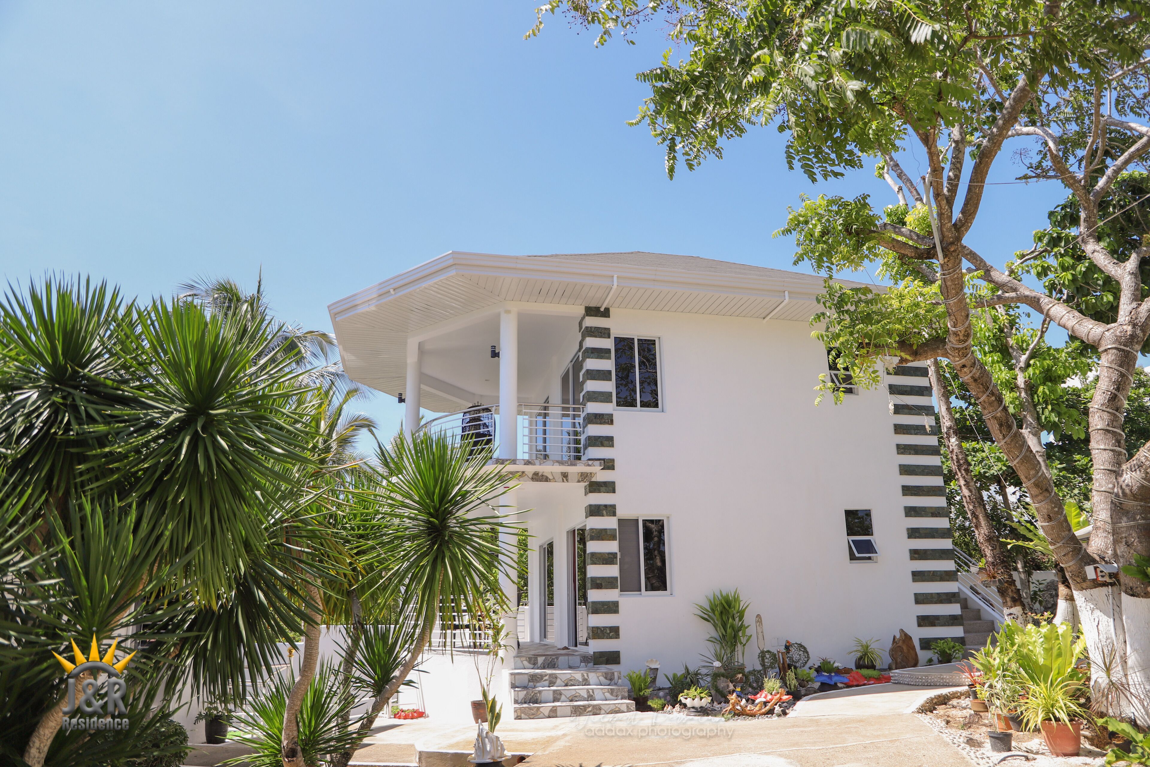 Une maison moderne à deux étages, avec une façade blanche, un porche abrité et un jardin aménagé comprenant diverses plantes et un palmier.