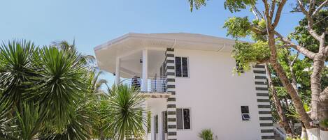A modern two-story house with a white exterior, a covered porch, and a landscaped garden with various plants and a palm tree.