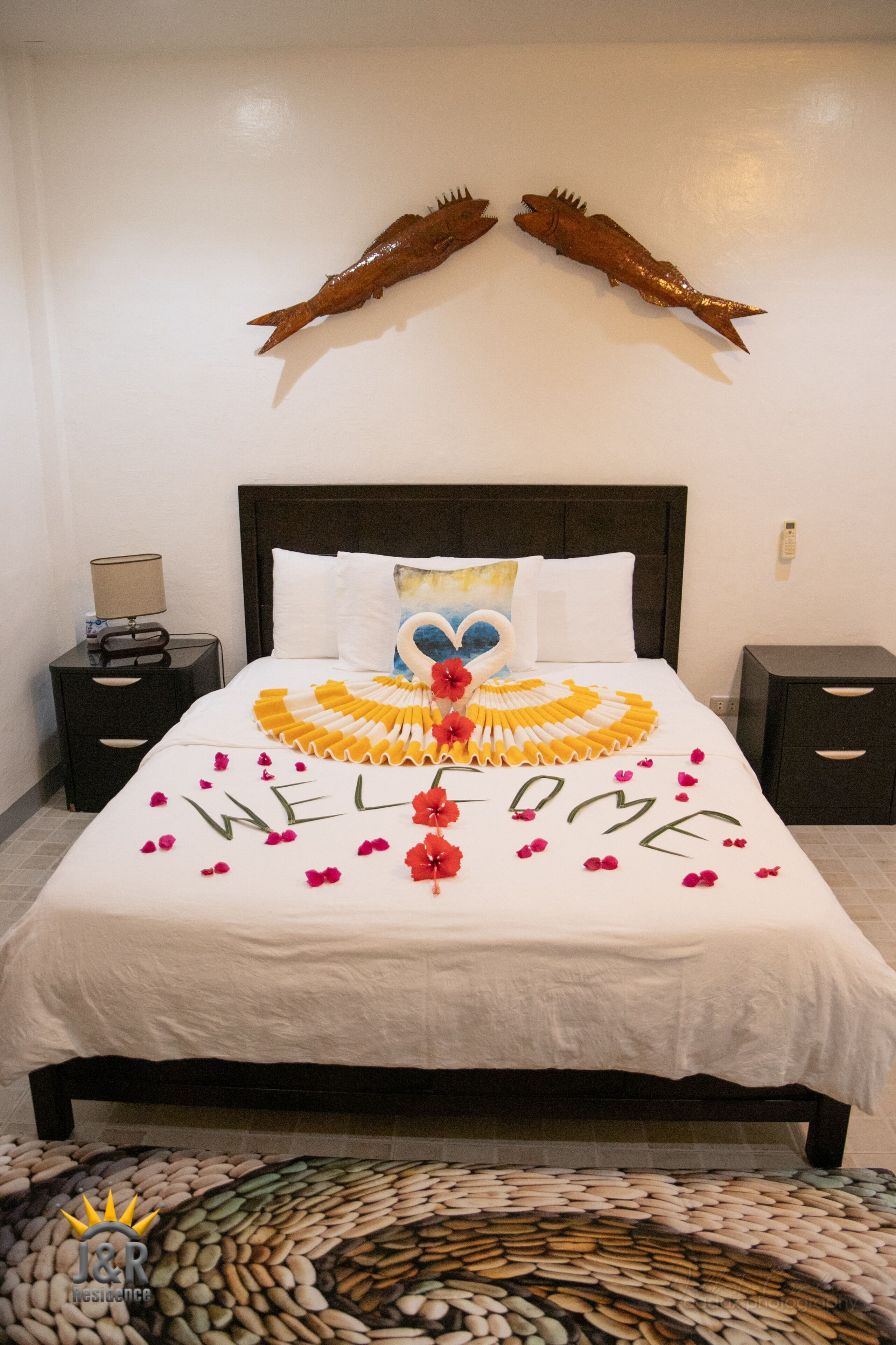 A neatly made bed with a welcome mat, decorative fish, and a heart-shaped pillow.