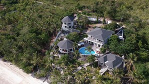 An aerial view of a large, multi-story house with a swimming pool, surrounded by dense tropical vegetation and a beach. - J&R Residence (Anda)