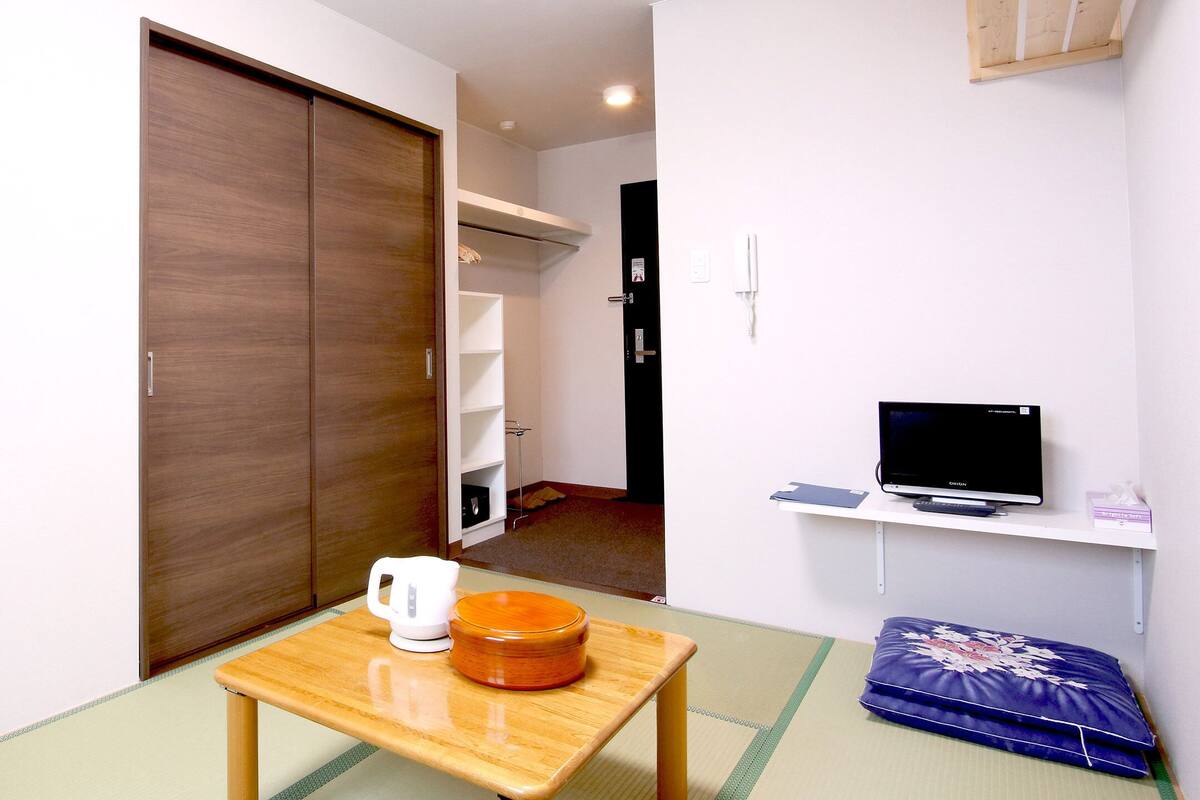 A room with a sliding door, a small table with a kettle, a television on a shelf, and a futon bed.