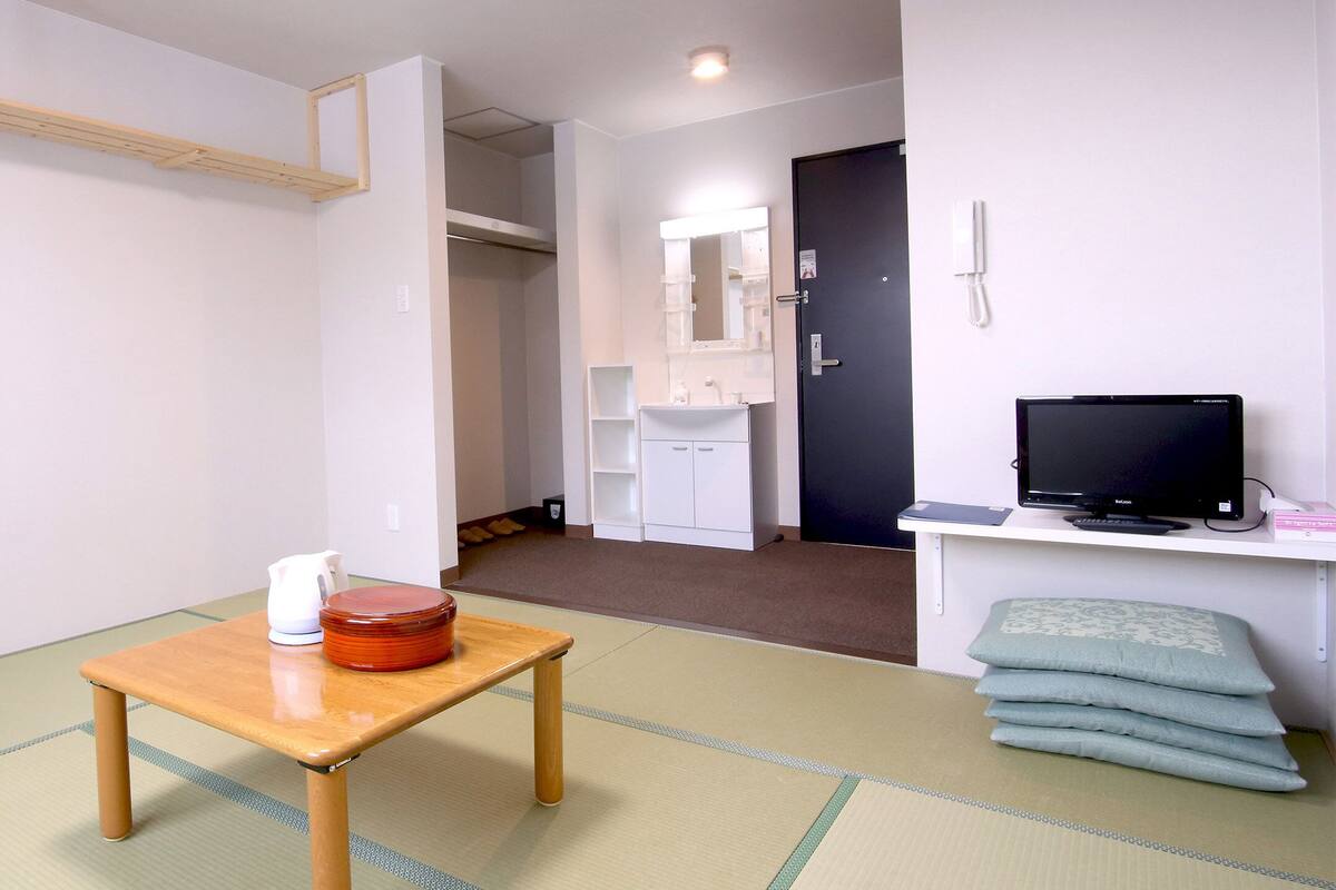 A room with tatami flooring, a wooden table with a kettle and a red container, a television on a stand, and a stack of pillows.