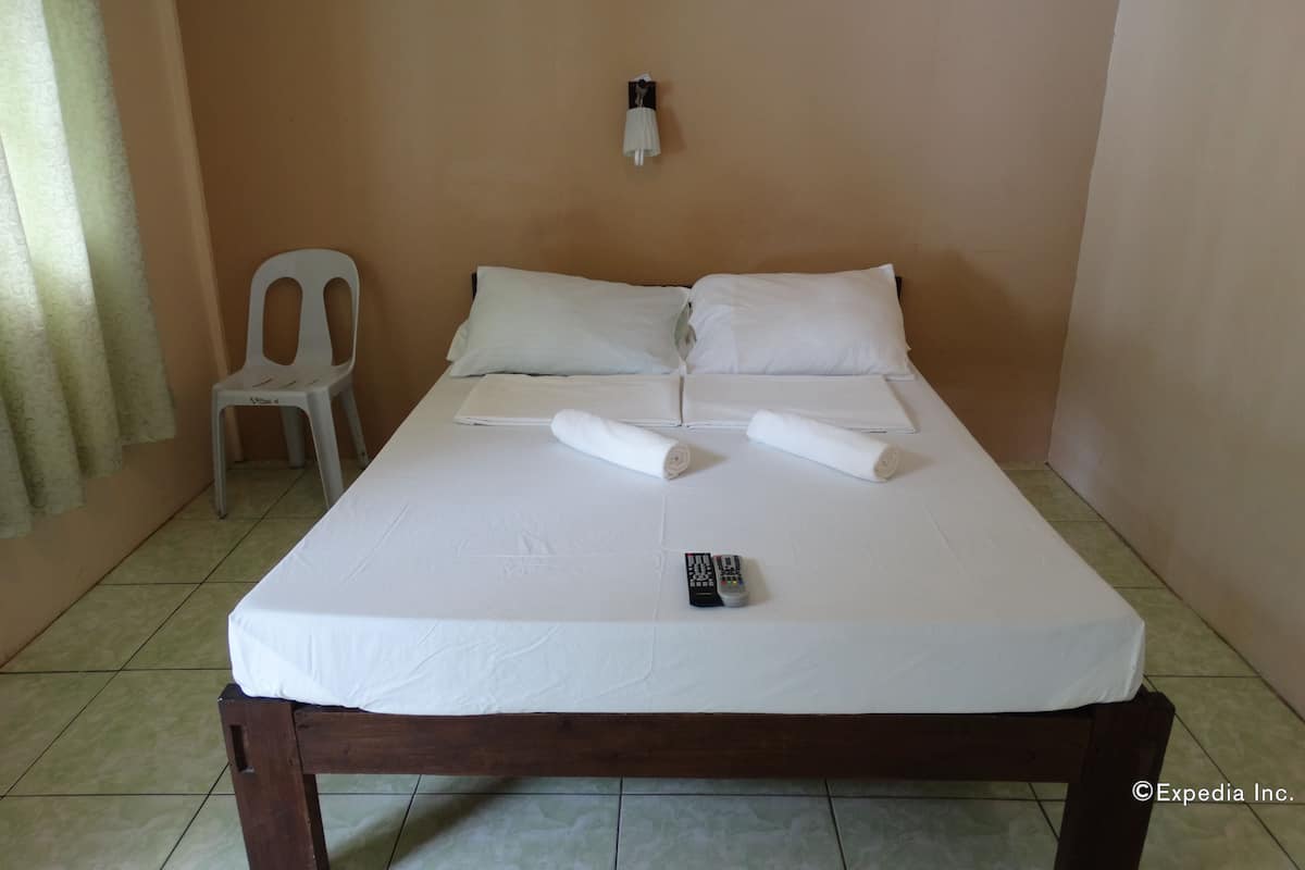 Un llit ben fet amb llençols blancs, un comandament a distància i una tovallola doblada sobre un brancal de fusta en una habitació amb parets beix i una cadira blanca.