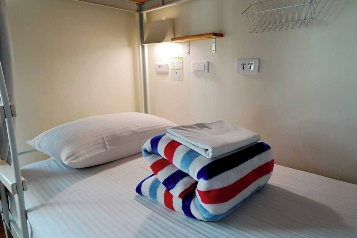Un lit superposé avec un oreiller blanc et une couverture à rayures. On peut voir des prises de courant et un interrupteur sur le mur.