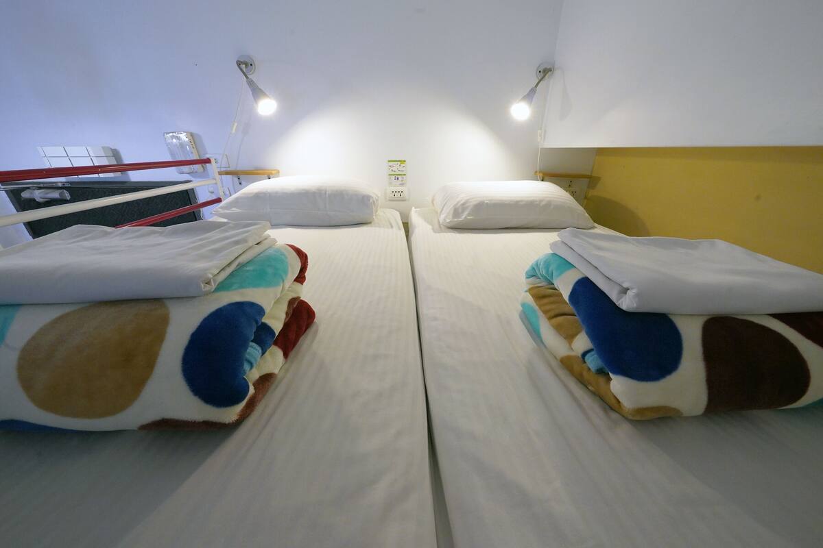 Une chambre d’hôtel avec deux lits, chacun recouvert d’une couverture et d’un oreiller à motifs, un mur blanc et un luminaire au plafond.