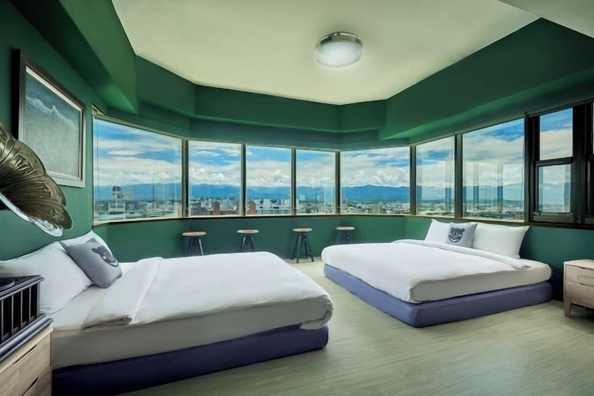 Una habitació d'hotel amb dos llits, grans finestres que donen vista a la ciutat i una làmpada al sostre.