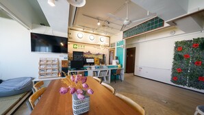 A modern interior with a wooden dining table, chairs, a vase with flowers, a flat-screen TV, a counter with a menu board, and a decorative wall with red tags. - An Lan Jie Hotel - Hostel (Chiayi City)
