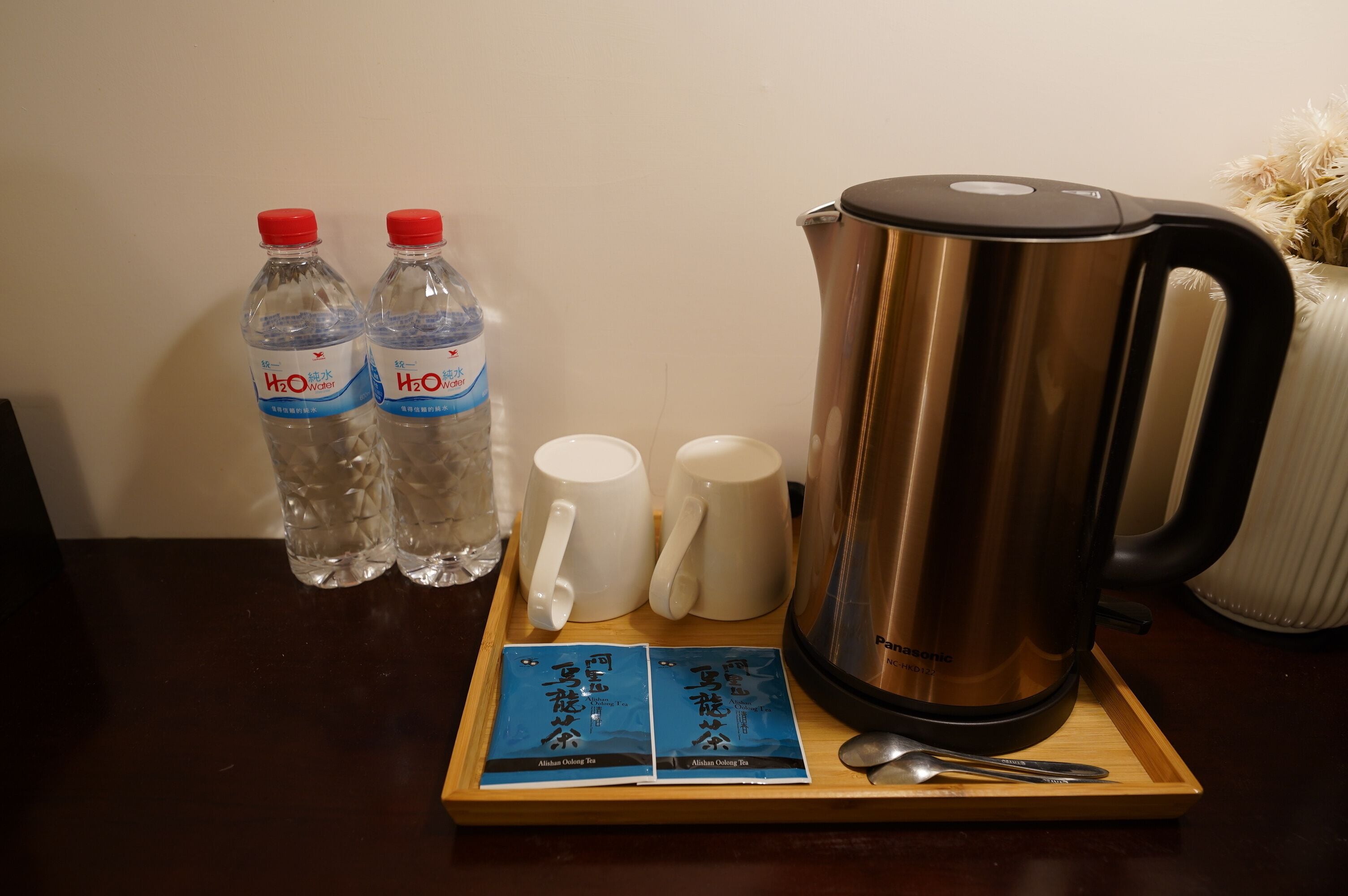 Una bandeja de madera con dos botellas de agua, dos tazas blancas, una tetera eléctrica de acero inoxidable y dos sobres de té.