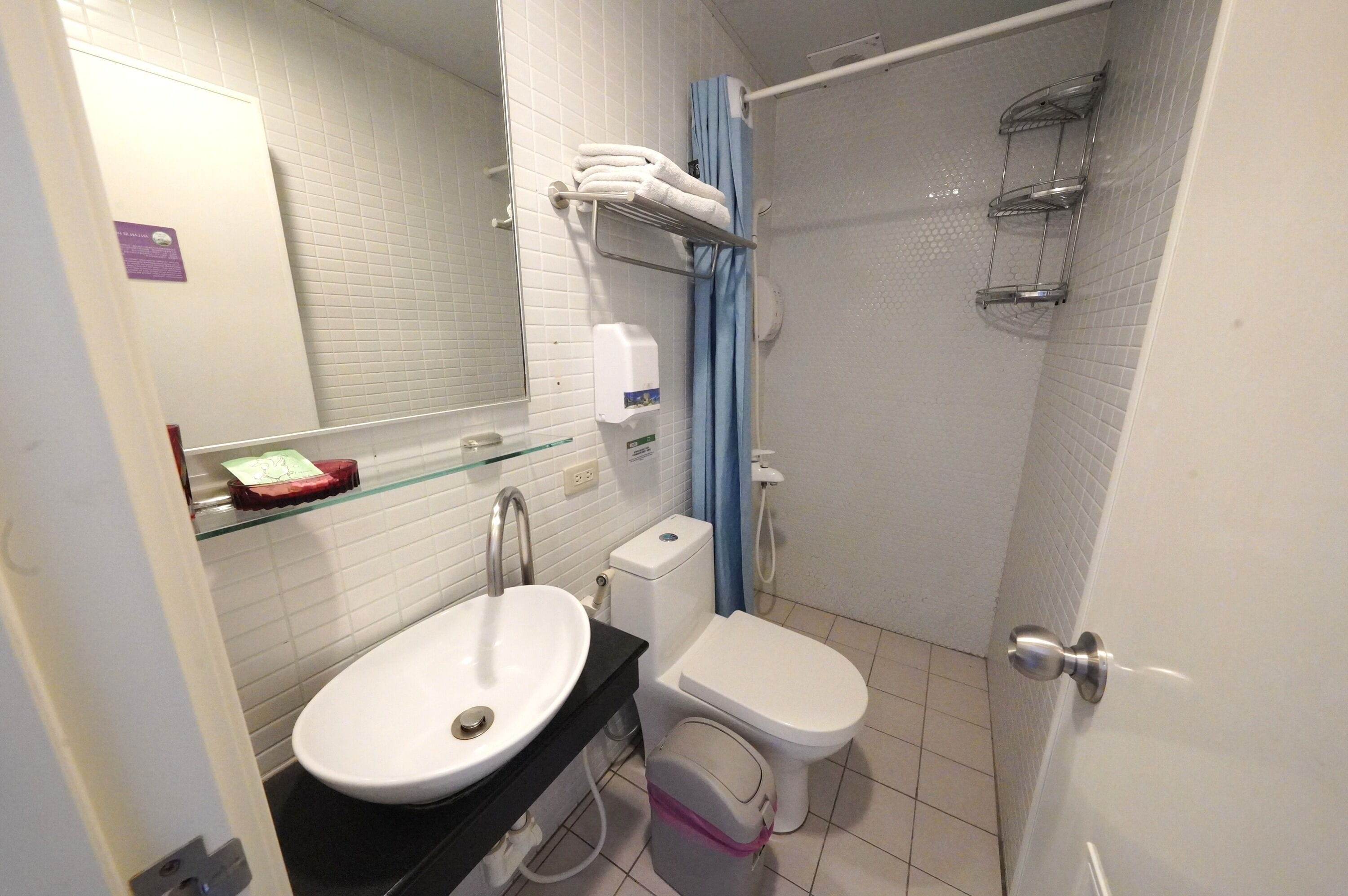 A bathroom with a white sink, toilet, and shower. There are towels on a rack and a soap dispenser on the wall.