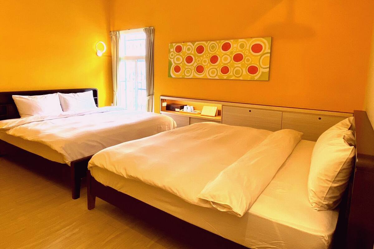 Une chambre d’hôtel avec deux lits, une fenêtre avec des rideaux, un tableau décoratif au mur et une table de chevet avec une lampe.