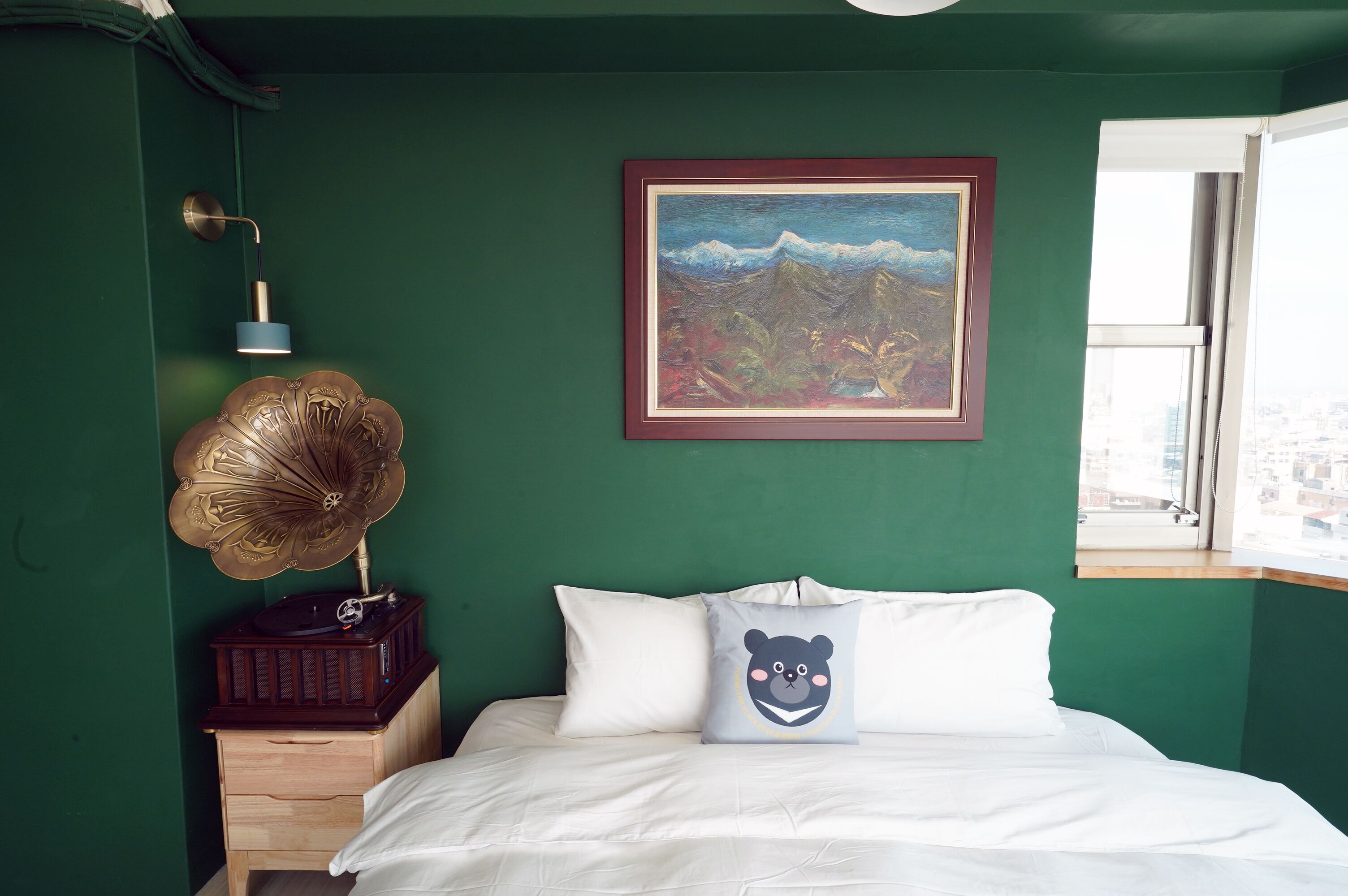 A bedroom with a green wall, a framed painting, a bed with white bedding, a pillow with a bear design, a vintage gramophone, and a bedside lamp.