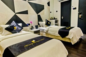 A hotel room with two beds, each with a black and white bedspread and matching pillows. There is a small table with a vase of flowers and a nightstand with a lamp. The room has a geometric patterned wall and a door with a cross on it.