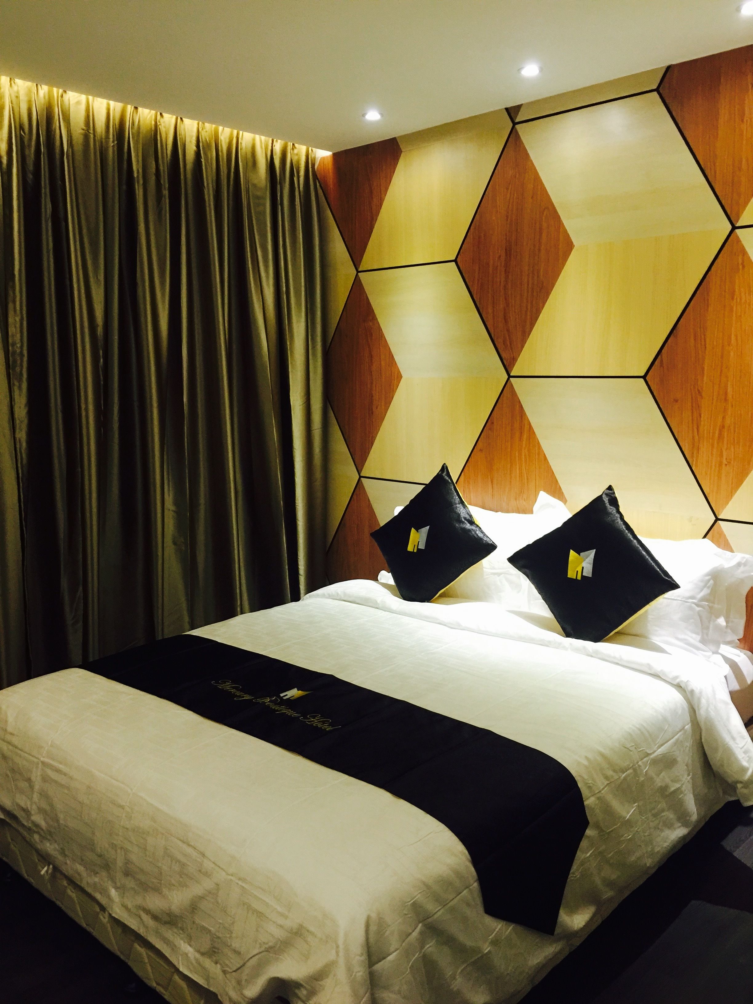 Habitación de hotel con cama, colcha blanco y negro, dos almohadas negras con un logotipo, cortina dorada y diseño geométrico en la pared.
