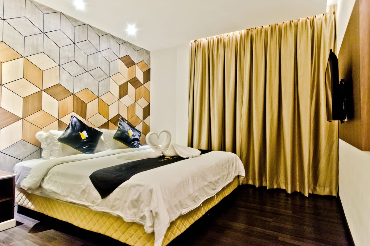 Une chambre d’hôtel dotée d’un décor mural géométrique moderne, d’un lit soigneusement fait avec une literie blanche et des coussins décoratifs, et d’une grande fenêtre ornée de rideaux dorés.