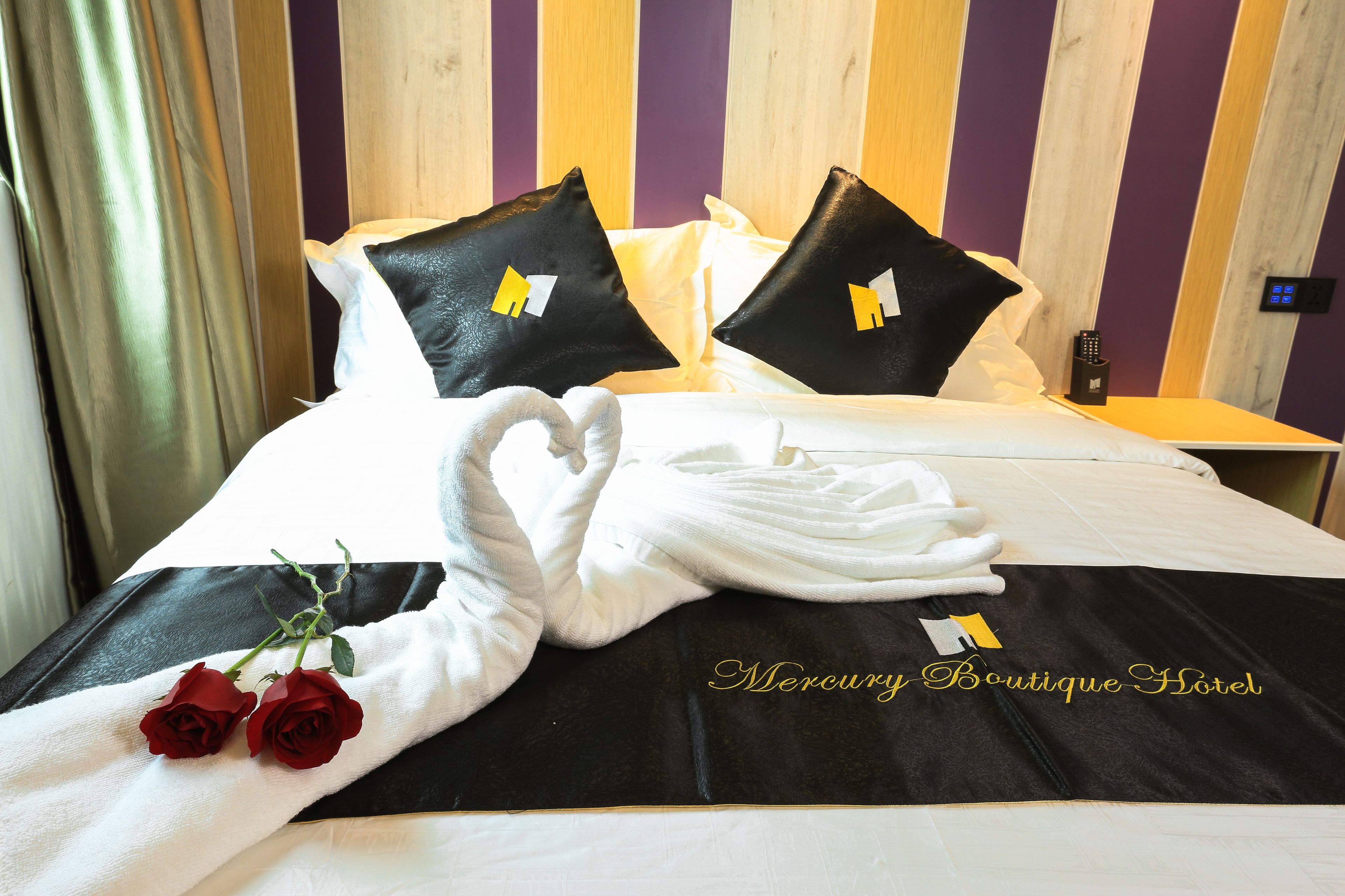 Quarto de hotel com uma cama, almofadas brancas e pretas com um logotipo, uma toalha branca dobrada e duas rosas vermelhas.