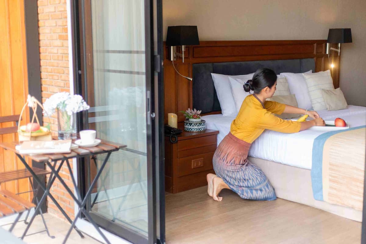 A hotel room with a bed, a wooden nightstand, a table with a vase and a cup, and a view through a glass door.