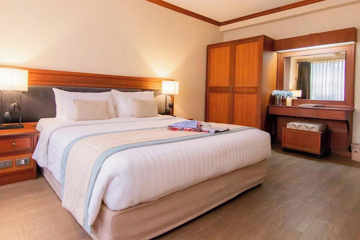 A hotel room with a large bed, wooden headboard, bedside lamps, a nightstand, a dresser, a mirror, and a small bench.