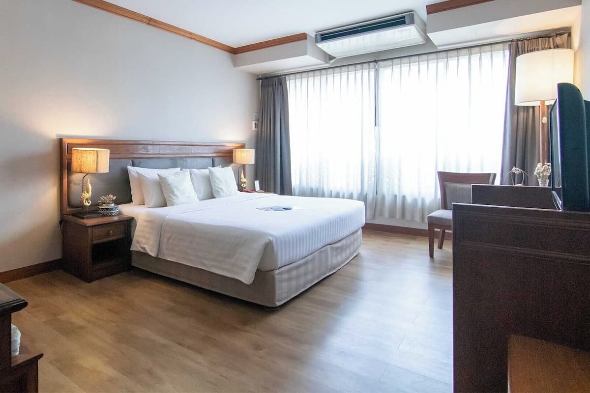 A hotel room with a large bed, a nightstand with a lamp, a desk with a chair, and a window with curtains.